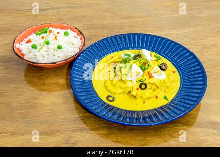 aji de gallina with boiled eggs and rice garnish with peas served in blue plate and bowl of white rice on oak wood table Stock Photo