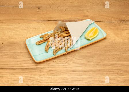 paper cone with fried anchovies on blue plate and lemon wedge on wooden table Stock Photo