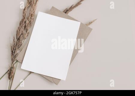 Fall wedding, birthday stationery composition. Blank greeting card, invitation mockup with brown envelope. Dry grass, festuca plant on Beige table Stock Photo