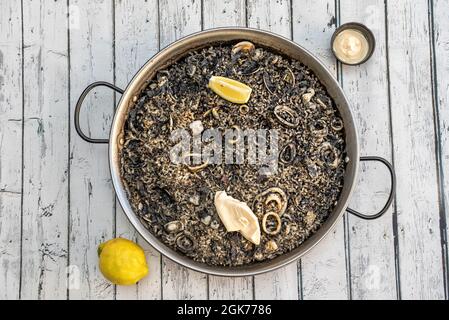 Spanish paella of black rice with squid, fish and clams with aioli sauce and lemon on white table Stock Photo