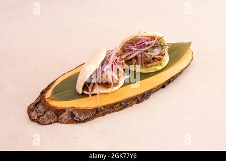 Two bao bread sandwiches stuffed with pork loin with lots of red onion presented on a banana leaf on top of a wooden board Stock Photo