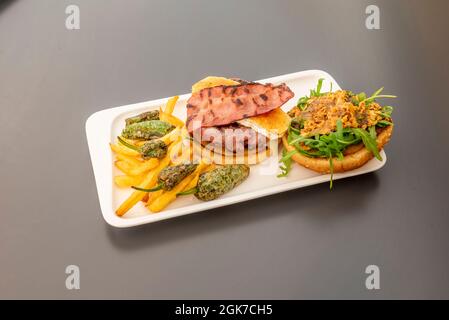 Gourmet beef burger with goat cheese, fried bacon with crispy onion with arugula and fried potatoes with padron peppers Stock Photo