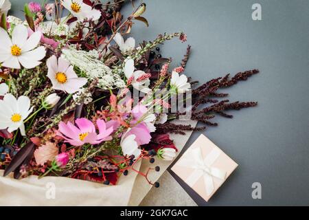 Fresh bouquet of white pink flowers with burgundy foliage wrapped in paper and arranged on grey background with gift box. Present for holiday Stock Photo