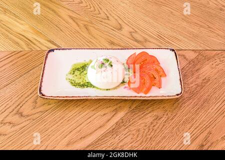 Burrata cheese with tomato slices, pesto sauce, olive oil and green sprouts on a white rectangular plate Stock Photo