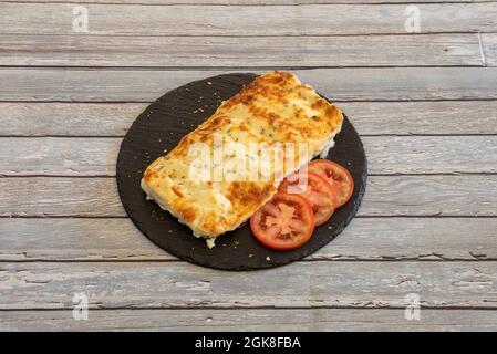 Portion of cannelloni stuffed with beef with tomato slices, gratin béchamel and oregano on a black slate plate Stock Photo