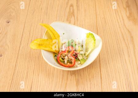 Delicious Peruvian marinated sea bass ceviche with banana chips, red pepper and ripe avocado on a white bowl Stock Photo