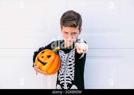 Serious boy in black skeleton costume holding Halloween Jack O Lantern pumpkin and pointing at camera while standing against white background Stock Photo