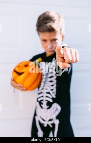 Serious boy in black skeleton costume holding Halloween Jack O Lantern pumpkin and pointing at camera while standing against white background Stock Photo