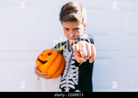 Serious boy in black skeleton costume holding Halloween Jack O Lantern pumpkin and pointing at camera while standing against white background Stock Photo