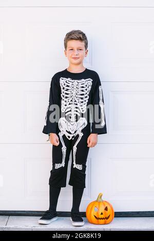 Full body of smiling preteen boy wearing black Halloween costume with skeleton print standing near carved Jack O Lantern pumpkin against white wall Stock Photo