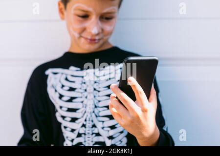Cheerful boy in black skeleton costume with painted face using on mobile phone while sitting near white wall on street Stock Photo