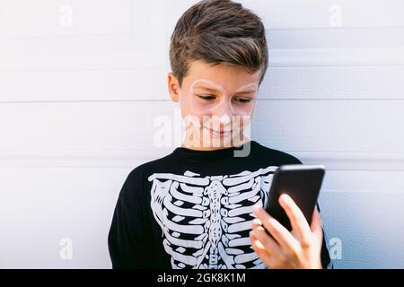 Cheerful boy in black skeleton costume with painted face using on mobile phone while sitting near white wall on street Stock Photo
