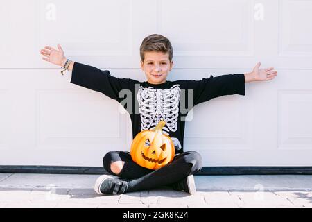 Full body of excited boy in skeleton costume with makeup and carved Halloween pumpkin raising arms and screaming with scary face while sitting near wh Stock Photo