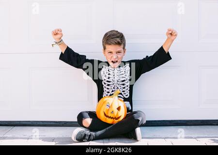 Full body of excited boy in skeleton costume with makeup and carved Halloween pumpkin raising arms and screaming with scary face while sitting near wh Stock Photo