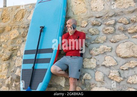 Male surfer in wetsuit leaning on stone wall speaking on smartphone with paddle and SUP board while preparing to surf on seashore Stock Photo