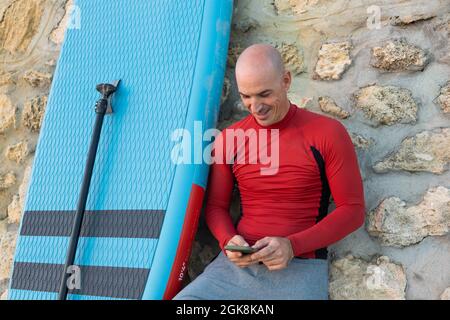 Male surfer in wetsuit leaning on stone wall browsing on smartphone with paddle and SUP board while preparing to surf on seashore Stock Photo