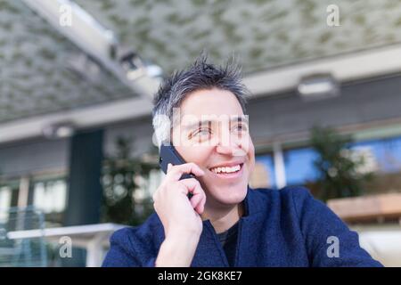 Happy young man in casual clothes smiling while sitting on bench and answering phone call Stock Photo