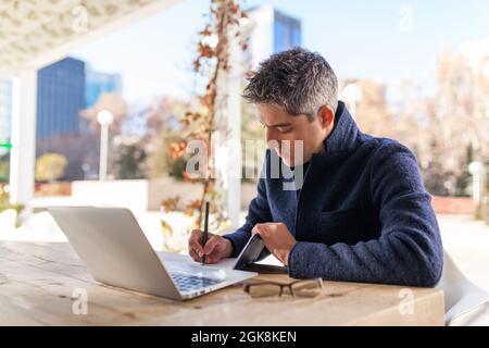 Side view of concentrated young bearded male freelancer in casual clothes and eyeglasses sitting at table with laptop and smartphone while working on Stock Photo