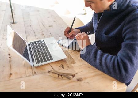 Side view of concentrated young bearded male freelancer in casual clothes and eyeglasses sitting at table with laptop and smartphone while working on Stock Photo