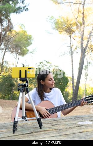 Cheerful female teen blogger playing acoustic guitar while recording video on cellphone on tripod in park Stock Photo