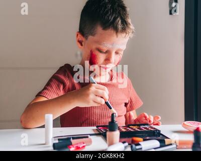 Charming child with makeup applicator looking away at table with eyeshadow palette Stock Photo