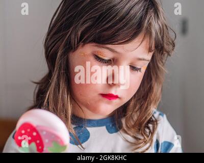 Kid with mirror making up face at table with assorted cosmetic products in house Stock Photo