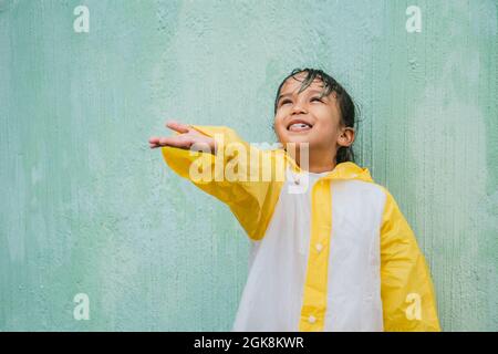 Charming ethnic child in slicker with palm up catching rain while looking up on pastel background Stock Photo