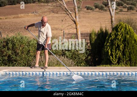 Full body of elderly male in eyeglasses cleaning water in swimming pool with mop in backyard Stock Photo