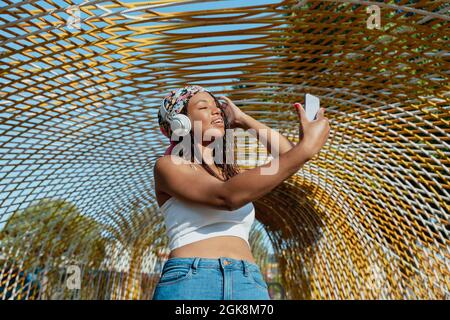 Positive young ethnic female with cellphone taking a selfie while listening to music from wireless headphones while laughing in town Stock Photo