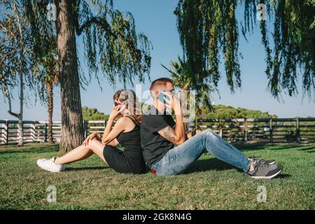 Side view of tattooed man with girlfriend in sunglasses talking on cellphones while sitting back to back on meadow in summer park Stock Photo