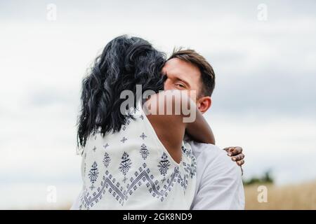 Happy man embracing Indian girlfriend standing in field under cloudy sky Stock Photo
