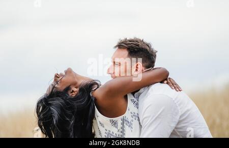Side view of smiling man embracing Indian girlfriend standing in field under cloudy sky Stock Photo