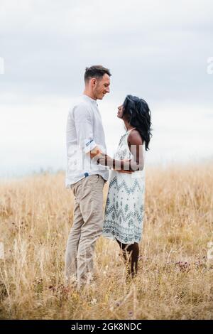 Side view of smiling man embracing Indian girlfriend while looking at each other in field under cloudy sky Stock Photo