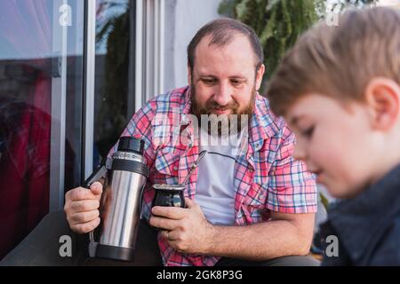 Hipster dad pouring herbal tea from thermos into calabash gourd against boy  with lumber on boardwalk Stock Photo - Alamy