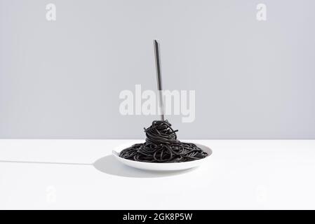 Stainless fork in full bowl of black spaghetti with cuttlefish ink on white table in studio on gray background Stock Photo