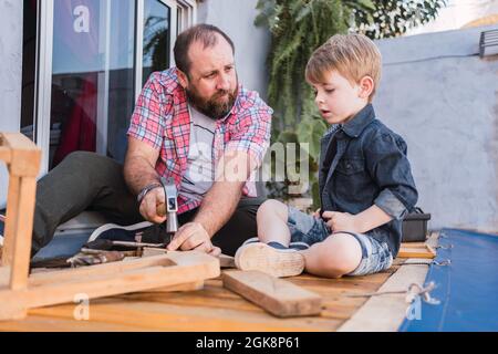 Bearded dad teaching son with hammer working with wood while sitting on boardwalk on weekend Stock Photo