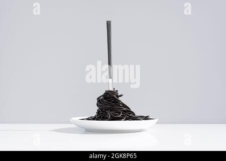 Stainless fork in full bowl of black spaghetti with cuttlefish ink on white table in studio on gray background Stock Photo
