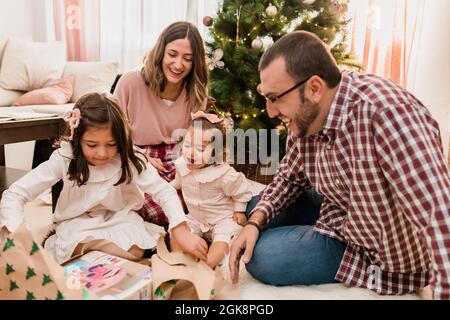 Surprised girls with cheerful parents opening gift box on floor while celebrating Christmas Day in house room Stock Photo