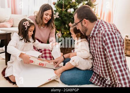 Surprised girls with cheerful parents opening gift box on floor while celebrating Christmas Day in house room Stock Photo