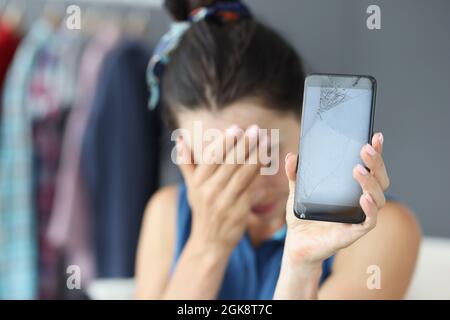 Sad woman covering her eyes with hand and holding broken mobile phone closeup Stock Photo
