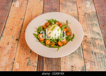 Burrata cheese salad with oil sauce with parsley and split cherry tomatoes on arugula and inside a white plate. Stock Photo