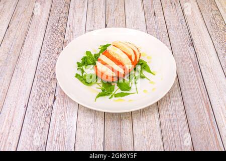Chopped tomato with burrata slices interspersed in a caprese salad with olive oil and arugula on a white plate Stock Photo
