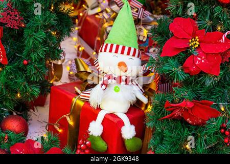Bright colorful toy snowman on wrapped gift holiday boxes on green fir tree branches background. Christmas and New Year celebration concept. Stock Photo