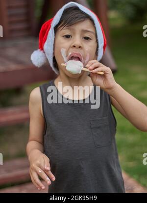 Brunette Little boy savoring an ice cream at Christmas time wearing a red Santa Claus hat and looking at camera. vertical. Argentina Stock Photo