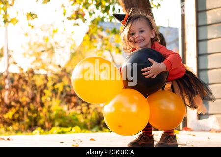 A cute little girl 2-3 in an orange and black Halloween witch costume with balloons, standing on the terrace of a wooden gray house Stock Photo