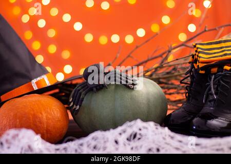 Hat, boots, stockings of a witch with branches, pumpkins and a spider on an orange background with bokeh. Halloween. Copy space. Stock Photo