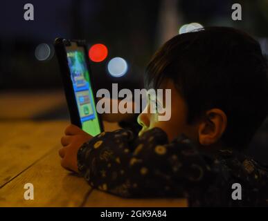 brunette little boy in a bar watching a video on a smart phone leaning on a wooden table, horizontal Stock Photo