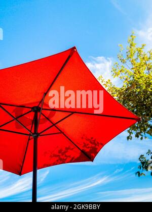 Underneath of bright red patio umbrella on a sunny blue sky day with next to tree foliage. Stock Photo