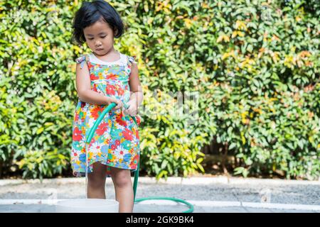 Shallow focus of a cute Southeast Asian kid holding a water hose in the sunny garden Stock Photo