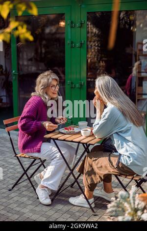 Joyful mature woman with friend small table with dessert and drinks outdoors Stock Photo